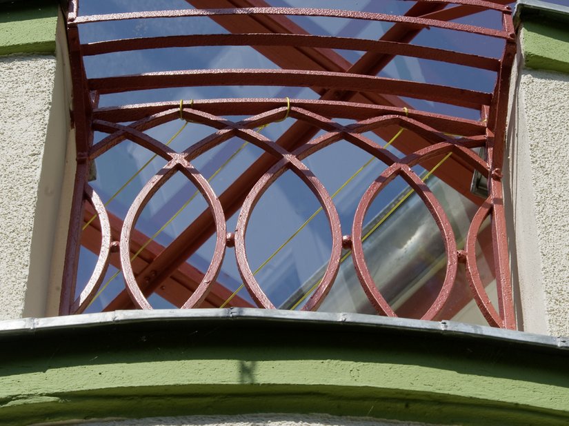 Ein rotes Metallgeländer rundet mit der Tragekonstruktion des Glasdaches über dem halbrunden Vorbau des Vordachs ab.
