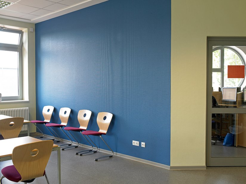 Die blaue Wand ist der „Ruhepol“. Buchefarbene Tische und Stühle werten den Arbeitsplatz auf, und die stoffbezogenen Sitzflächen in angenehmem Rotviolett laden zum Verweilen ein.