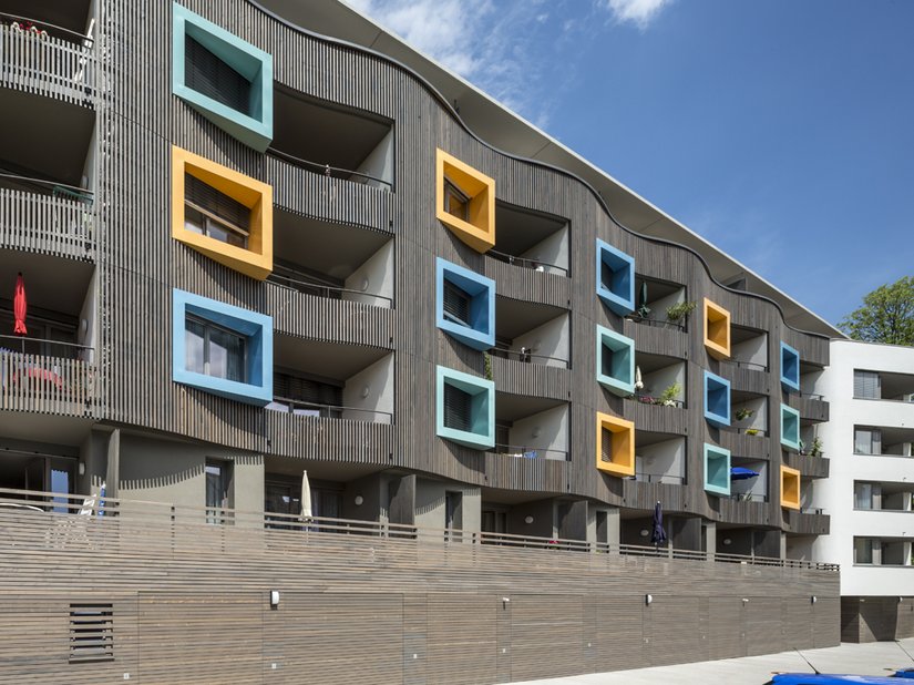 Die Fassade wird hier zu einer gewellten Haut aus Holz, in der Balkone und farbige Guckkästen der einzelnen Wohnungen integriert sind.