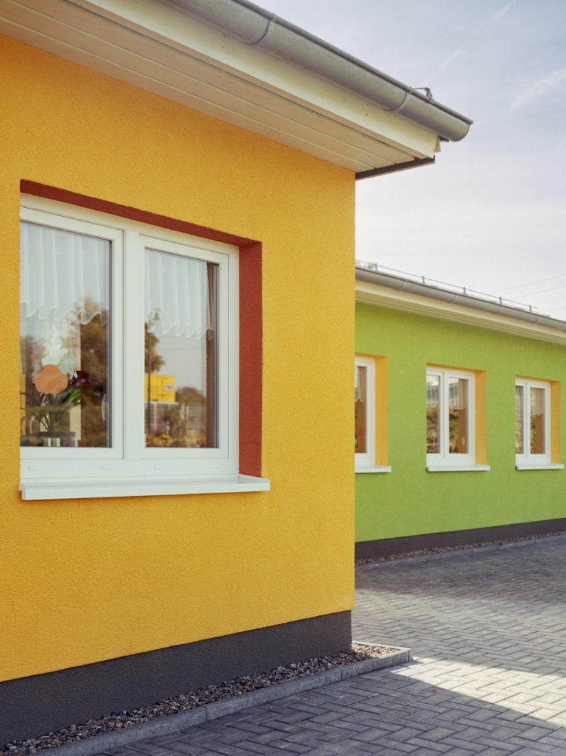 Die Wandfarbe des linken ziert die Innenseite der Fensteraussparungen des rechts liegenden Gebäudes, sodass ein Zusammenspiel der Farben entsteht.